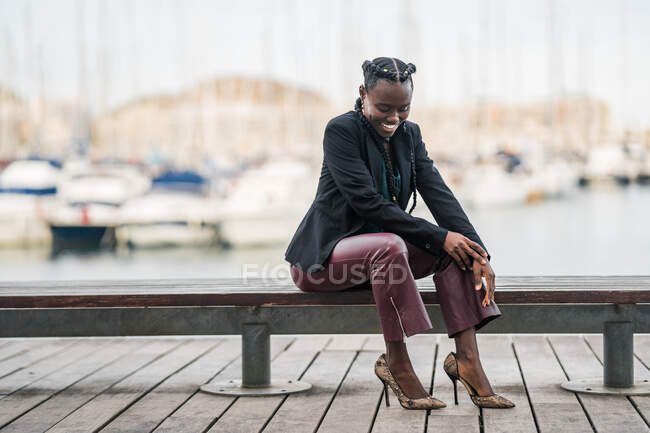 Stylish trendy smiling African American lady spending time sitting on wooden low bench in park in bright day — Stock Photo