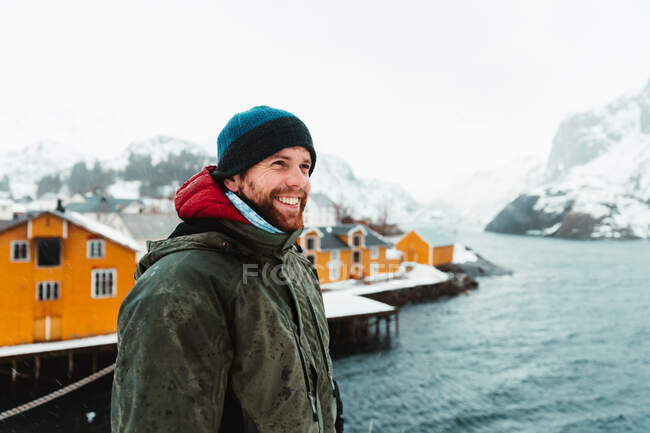 Вид сбоку человека в верхней одежде, стоящего на берегу моря на фоне желтых домов и заснеженного горного хребта в зимний день на Лофских островах, Норвегия — стоковое фото