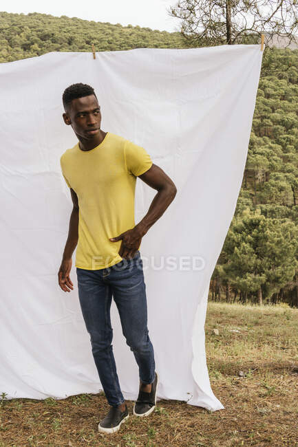 Серьёзный афроамериканец, стоящий на фоне белого натурального текстиля, стоящего у бельевой верёвки в природе — стоковое фото
