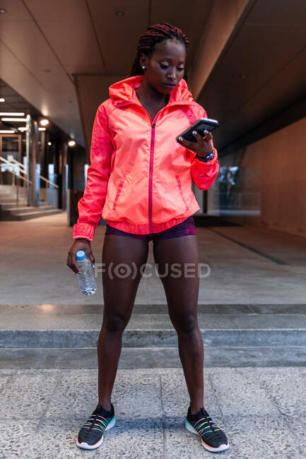 Спортивна афроамериканська спортсменка в червоній куртці стоїть на вулиці і користується смартфоном. — стокове фото