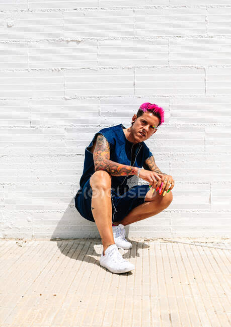Selbstbewusster queerer Mann mit bunten Nägeln und rosa Haaren, der im Sommer auf der Straße sitzt und in die Kamera blickt — Stockfoto