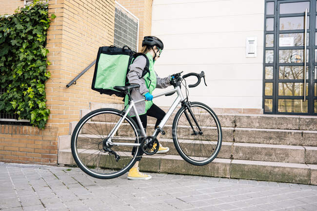 Full body female courier with thermal bag carrying bike on stairs towards building entrance while making delivery — Stock Photo