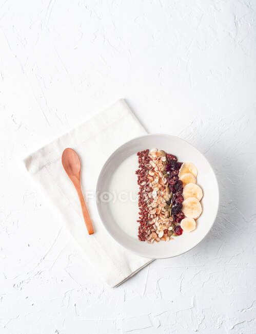 Vista superior del tazón con yogur blanco cubierto con cereales variados y plátano en rodajas servido para un desayuno saludable en la mesa con cuchara de madera - foto de stock