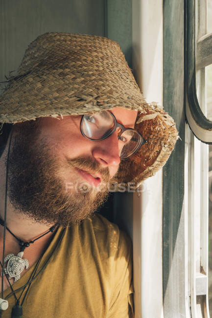 Smiling male hipster in straw hat traveling by train in summer and looking out of window — Stock Photo