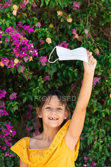 Zufriedenes Kind mit erhobenem Arm zeigt medizinische Maske beim Blick in die Kamera gegen blühende Pflanze im Garten — Stockfoto
