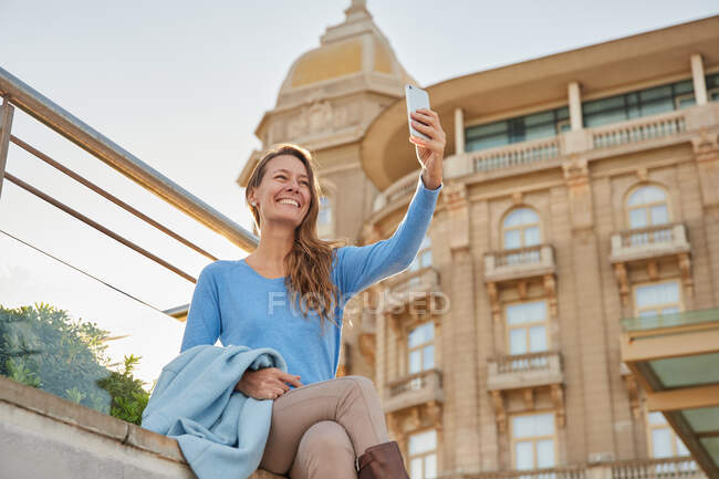 From below of smiling adult lady in casual clothes standing near fence and old building while taking selfie in city district in daylight — Stock Photo