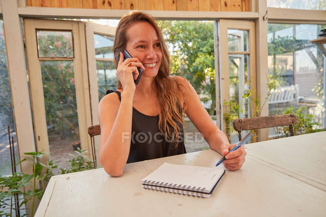 Smiling female gardener speaking on mobile phone and taking notes while sitting at table and working in hothouse in garden — Stock Photo