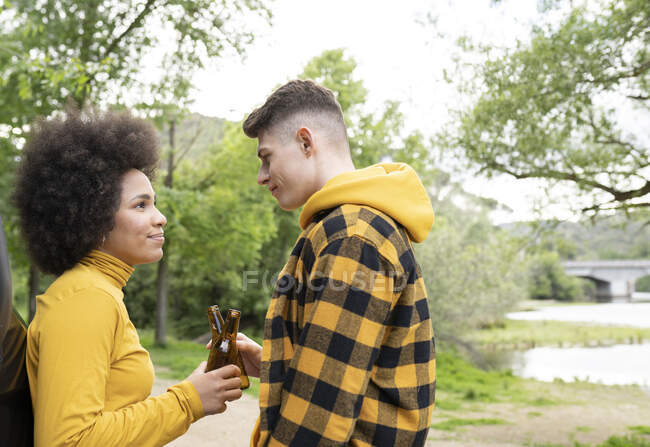 Vista lateral del joven multirracial y la mujer tintineo botellas de cerveza y mirándose mientras se relaja en la naturaleza en el día de verano - foto de stock