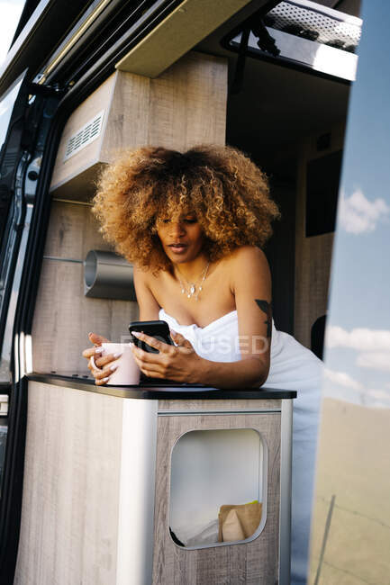 Glad femme afro-américaine avec tasse de boisson chaude souriant et téléphone portable de navigation tout en se reposant dans la caravane contemporaine le matin — Photo de stock
