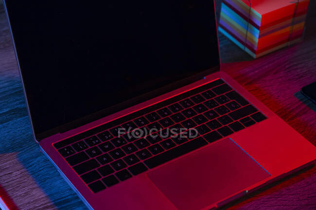 High angle of opened netbook with black screen placed on wooden table near note papers in dark workspace with neon lights — Stock Photo