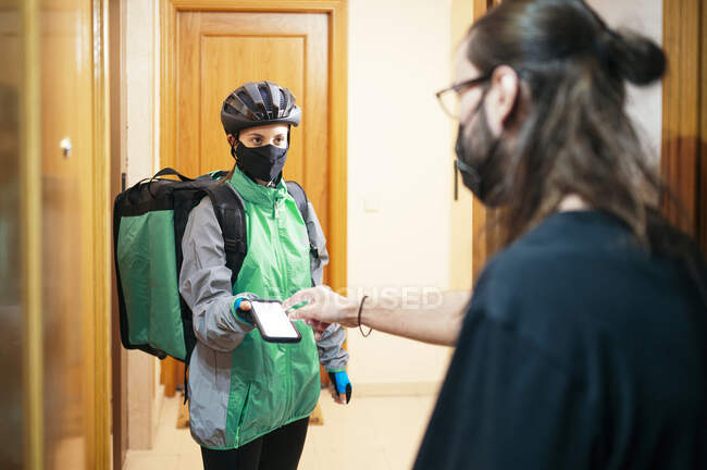 Male customer entering password on smartphone with blank screen while paying to delivery woman in mask during epidemic — Stock Photo