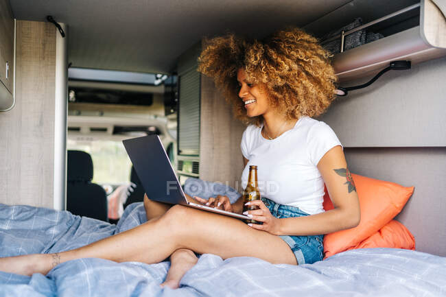 Jovem viajante afro-americana com cabelo encaracolado bebendo cerveja e assistindo filme no laptop enquanto descansa dentro van campista durante as férias de verão — Fotografia de Stock