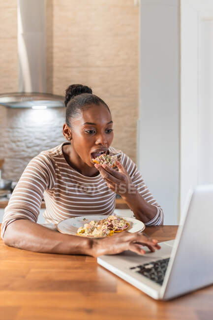 African American female enjoying tasty patacon with topping while surfing internet on netbook in kitchen at home — Stock Photo