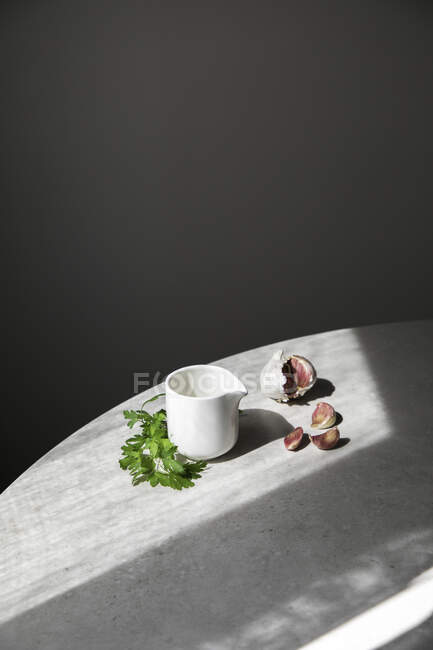 De cima dentes de alho e salsa verde fresca colocados na mesa redonda perto do barco de molho cerâmico no fundo cinza — Fotografia de Stock