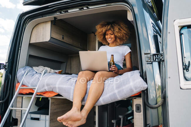 Jovem viajante afro-americana com cabelo encaracolado bebendo cerveja e assistindo filme no laptop enquanto descansa dentro van campista durante as férias de verão — Fotografia de Stock