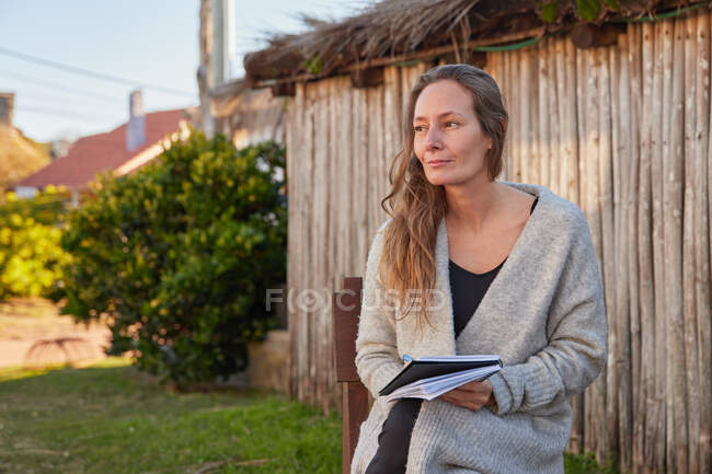 Боковой вид восхищенной женщины, пишущей в тетрадке, сидя на деревянном стуле на заднем дворе летом — стоковое фото
