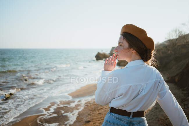 Обратный вид на мечтательную молодую путешественницу в стильном наряде и берете с видом на живописный морской пейзаж, стоящий на песчаном пляже в солнечный день — стоковое фото