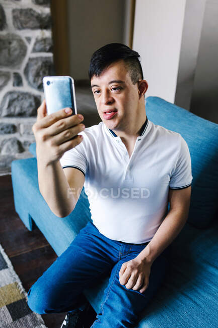 Alto ángulo de adolescente latino con síndrome de Down tomando auto disparo en el teléfono inteligente mientras está sentado en el sofá en casa - foto de stock