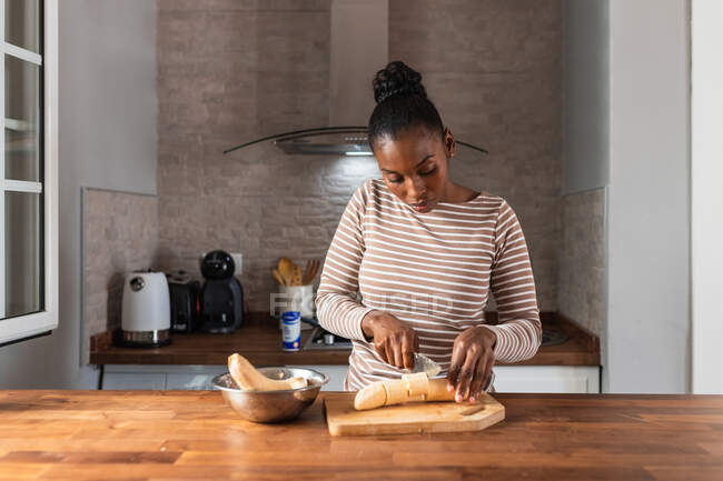 Donna nera in abbigliamento con ornamento a righe tagliando banane da cucina con coltello sul tagliere a tavola in casa — Foto stock