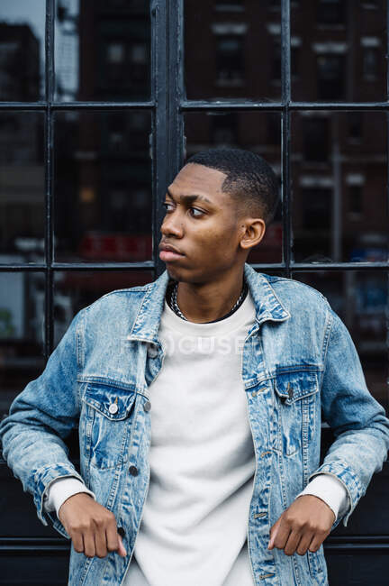 Black trendy serious male with silver chain on neck in blue denim jacket looking away on street — Stock Photo
