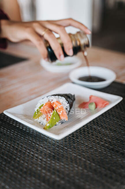 Coltiva una femmina irriconoscibile versando la salsa di soia nella ciotola contro il gustoso cono di sushi con fette di salmone e avocado sul piatto. — Foto stock