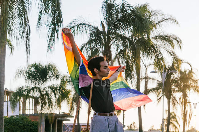 Contenido gay macho caminando con arco iris LGBT bandera en levantado manos a lo largo de calle en tropical ciudad - foto de stock
