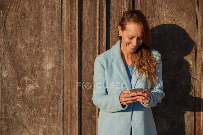 Happy adult woman in blue coat leaning on old building while browsing on cellphone in city district in sunny day — Stock Photo