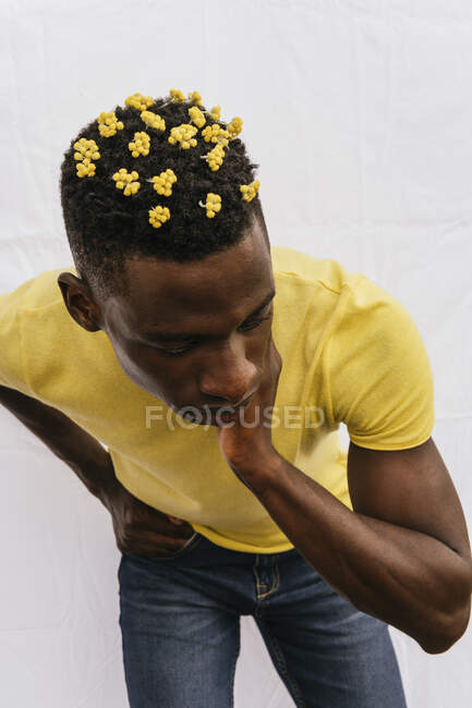 Beau mâle afro-américain avec des fleurs jaunes dans les cheveux touchant le cou et regardant vers le bas sur fond blanc — Photo de stock