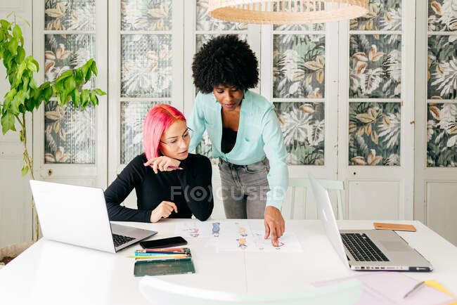 Jeunes créatrices de mode multiraciales discutant de croquis pour une nouvelle collection tout en travaillant ensemble à table dans une pièce lumineuse — Photo de stock
