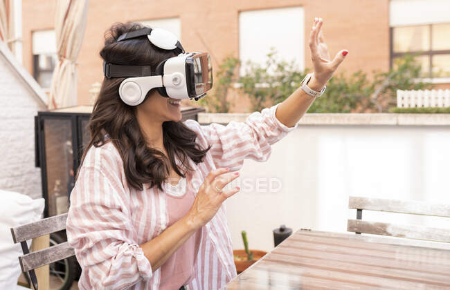 Visão lateral de uma mulher feliz em óculos de realidade virtual experimentando realidade virtual enquanto se senta à mesa no terraço — Fotografia de Stock