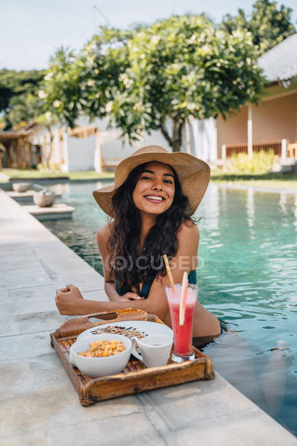 Cheerful female tourist leaning on poolside while looking at camera against tray with yummy breakfast in sunlight — Stock Photo