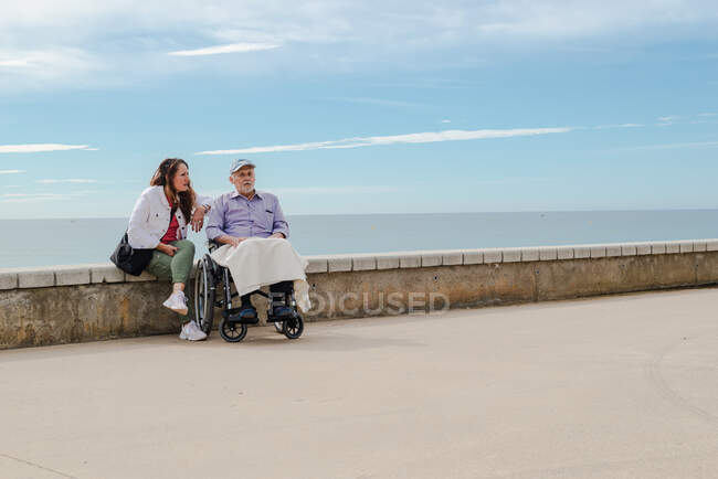 Доросла дочка і батько похилого віку в інвалідному візку, що голиться на набережній проти моря разом влітку — стокове фото