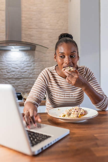 Afro-americano feminino desfrutando saboroso patacon com cobertura enquanto navega na internet no netbook na cozinha em casa — Fotografia de Stock