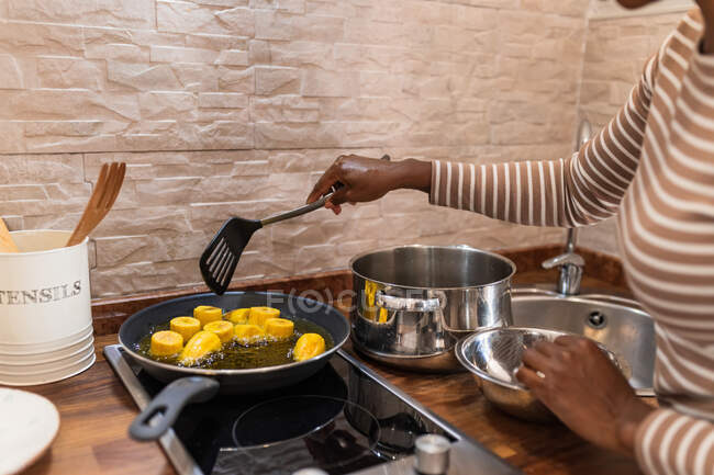Crop anonymous ethnic person frying cooking banana pieces in pan with hot oil on stove while preparing patacones in kitchen — Stock Photo