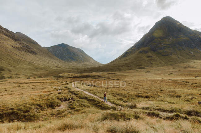 Visão traseira do homem irreconhecível andando no caminho da sujeira na encosta gramada áspera durante a viagem através de Glencoe no campo do Reino Unido no dia nublado — Fotografia de Stock