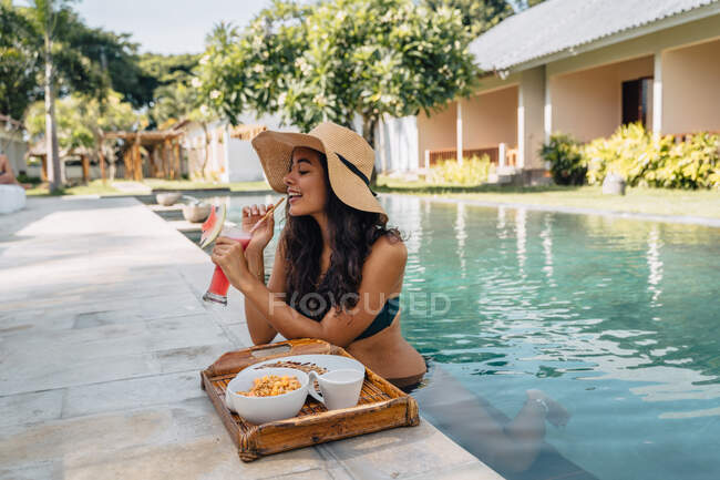 Весела жінка-туристка в купальнику насолоджується освіжаючим напоєм, спираючись на басейн зі смачним сніданком на підносі в тропічному курорті — стокове фото