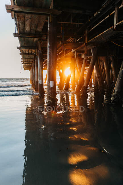 Luz solar brilhante à noite penetrando pilhas de cais de Santa Mônica com ondas pacíficas do oceano correndo na praia na Califórnia — Fotografia de Stock