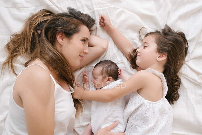 Сверху улыбающиеся мать и маленькая девочка лежат на кровати с симпатичным младенцем — стоковое фото