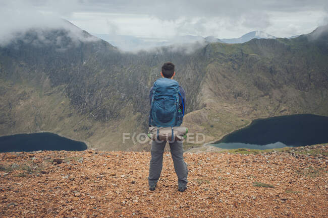 Visão traseira do viajante anônimo masculino com mochila em pé no miradouro nas terras altas e admirando paisagens incríveis da cordilheira durante o trekking no País de Gales no dia nublado — Fotografia de Stock