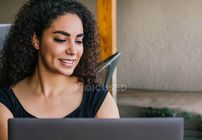 Zufriedene junge ethnische Freiberuflerin surft auf der Laptop-Tastatur, während sie an einem Remote-Projekt auf der Café-Terrasse arbeitet — Stockfoto