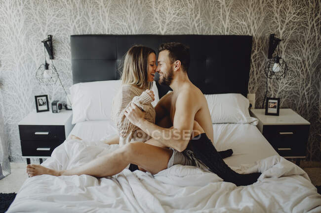 Alegre joven hombre y mujer sonriendo y abrazándose mientras están sentados en la cómoda cama en casa juntos - foto de stock