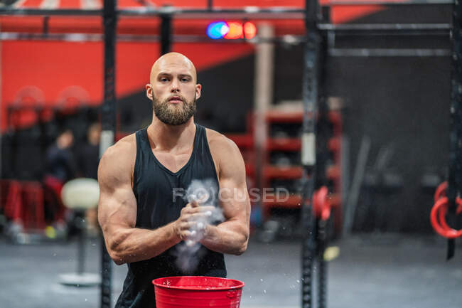 Muscular bearded guy in sportswear looking at camera clapping hands and spreading powder during bodybuilding workout in gym — Stock Photo