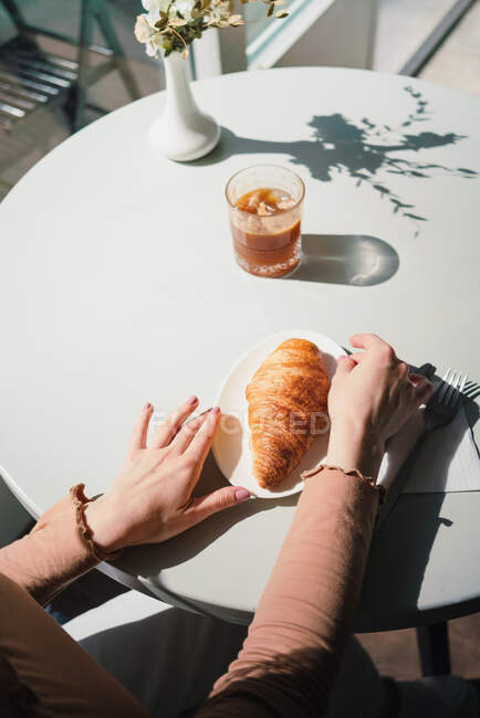 Anonymous female in beret sitting at table in cafe with aromatic glass of coffee and freshly baked croissant — Stock Photo