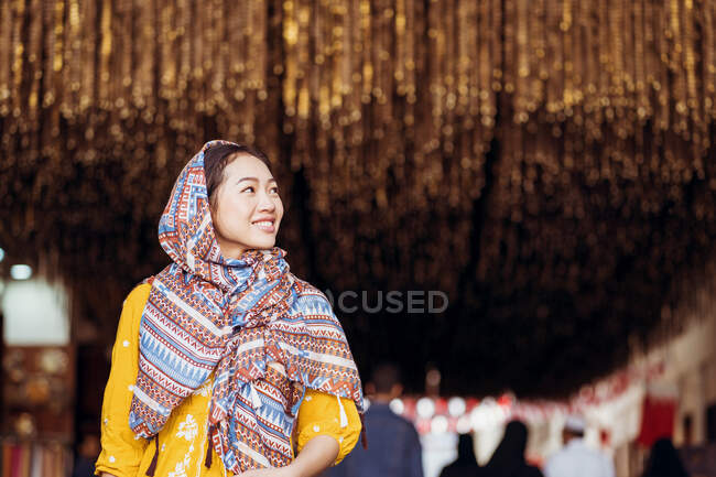 Jeune femme orientale en costume traditionnel et foulard souriant et regardant loin tout en se tenant debout sur un fond flou du vieux bazar souk Manama dans la ville de Manama au Bahreïn — Photo de stock
