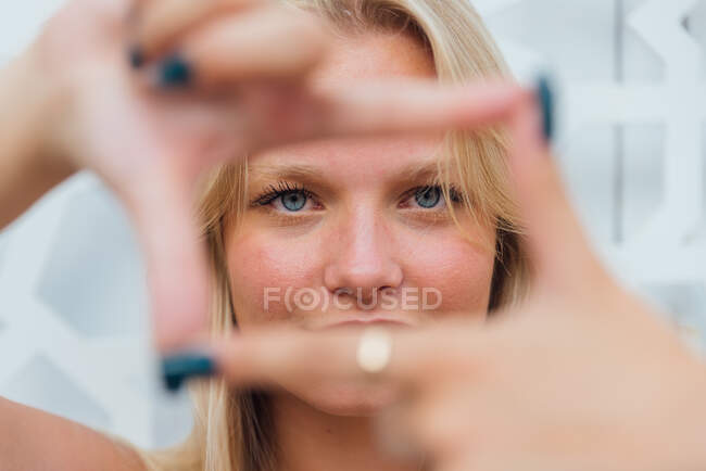 Zufriedene Frau mit blonden Haaren, die ein Rahmenzeichen zeigt und durch die Finger in die Kamera schaut — Stockfoto