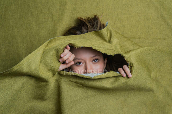 Молодая женщина с зелеными глазами смотрит в камеру, прячась за порванной зеленой тканью — стоковое фото