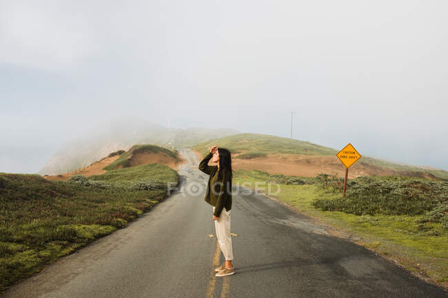 Comprimento total de mulher alegre de mãos dadas na cabeça em prazer da luz solar em pé na rota vazia do penhasco costeiro na Califórnia — Fotografia de Stock