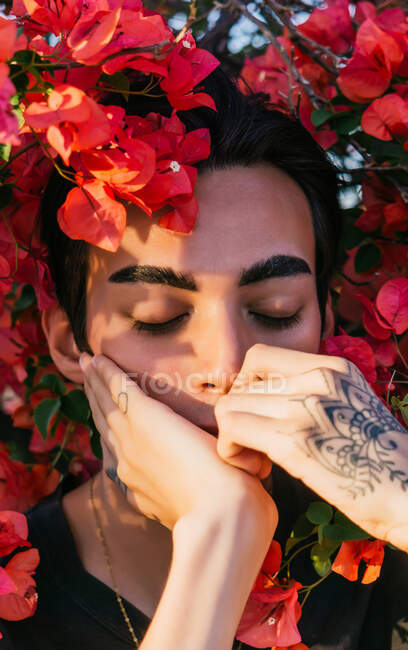 Serene gay mâle penché sur les mains avec tatouage avec les yeux fermés dans le parc d'été avec des fleurs florissantes — Photo de stock