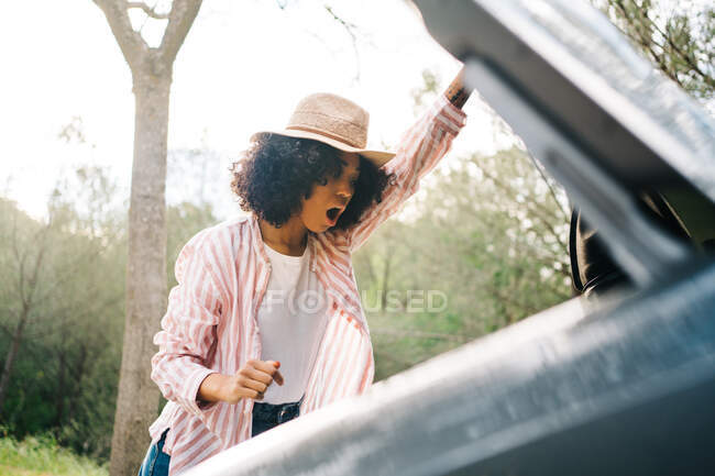 Side view of unhappy young African American female driver getting surprised while checking broken car during trip through forest — Stock Photo