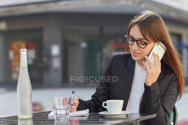 Positive young business lady in elegant suit and eyeglasses taking notes in notebook during phone conversation while sitting at table in outdoor cafe in city — Stock Photo
